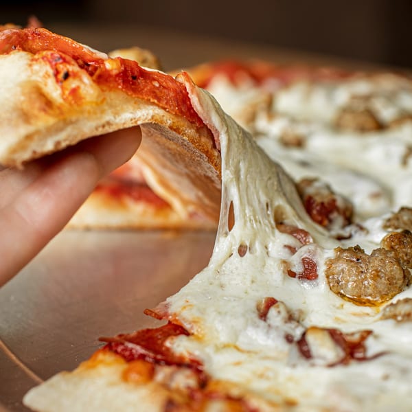 A hand lifts a slice of Hula Pizzeria & Grill pizza, stretching gooey cheese and revealing sausage.