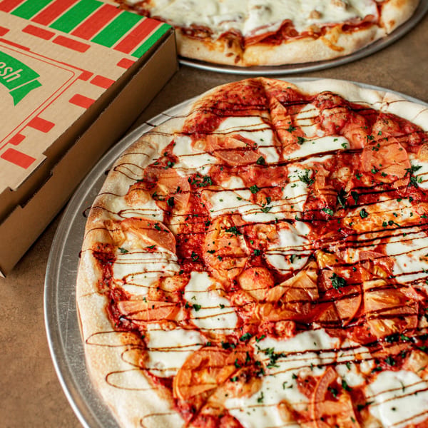Two Hula Pizzeria & Grill pizzas, one with tomatoes and balsamic glaze, next to a pizza box.