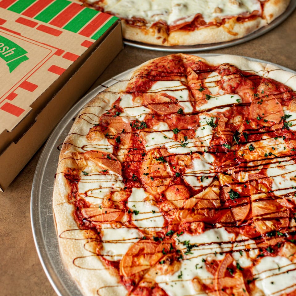 Two Hula Pizzeria & Grill pizzas, one with tomatoes and balsamic glaze, next to a pizza box.